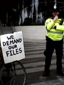 Police officer next to 'We Demand Our Files' placard
